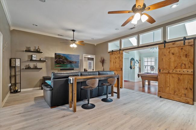 Living room featuring a barn door, ceiling fan, baseboards, light wood-type flooring, and ornamental molding | Image 7