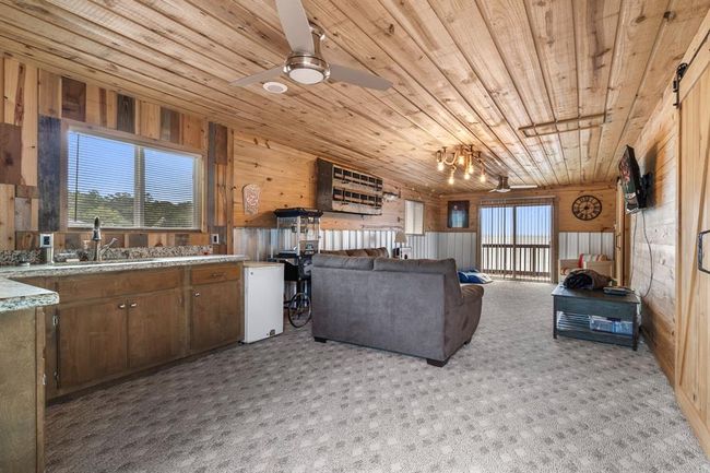 This cozy living space features rustic wood paneling on the walls and ceiling, a comfortable seating area, and large windows offering ample natural light. A compact kitchen with granite countertops and a mini-fridge is included, while sliding glass doors lead to a balcony with a scenic view. | Image 37