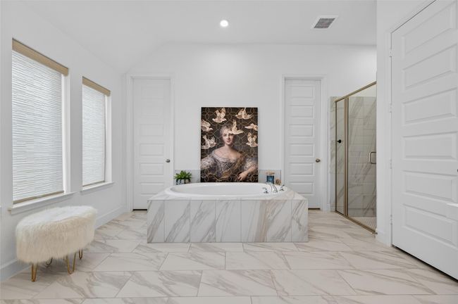 Elegant primary bath showcasing a marble-look soaking tub as the focal point, flanked by dual closet doors and a glass-enclosed shower. Expansive tile flooring and triple windows complete the spa-like ambiance with abundant natural light. | Image 27
