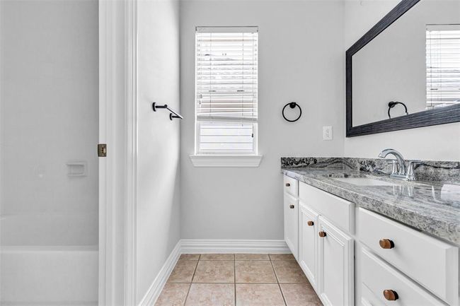 This bathroom features a sleek design with white cabinetry and a granite countertop. It includes a large mirror, a window for natural light, and a combination bathtub and shower. The neutral tile flooring adds a clean and modern touch. | Image 36