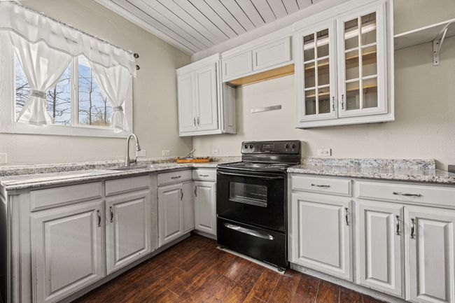 Kitchen with black range with electric cooktop, a sink, white cabinets, dark wood finished floors, and glass insert cabinets | Image 12