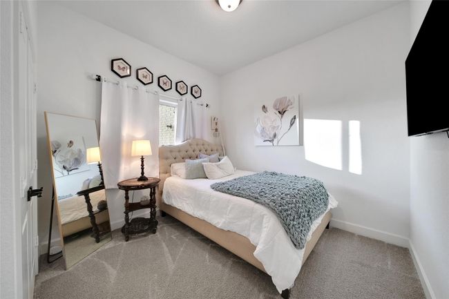 This cozy bedroom features a plush bed with a tufted headboard, soft carpeting, and a large wall-mounted TV. Natural light flows through a window with white curtains, complemented by elegant decor and a full-length mirror. | Image 36