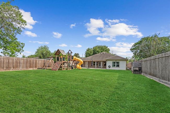 This image showcases a spacious backyard with a large, lush green lawn, a playset for children, and a privacy fence. The back of a single-story home is visible, indicating a family-friendly living space. | Image 37