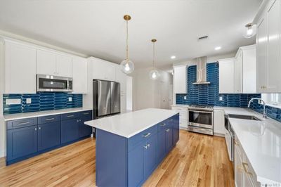 Kitchen with wall chimney range hood, light hardwood / wood-style flooring, tasteful backsplash, blue cabinetry, and stainless steel appliances | Image 3