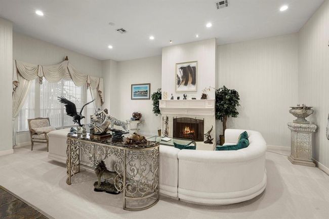 This is an elegantly furnished living room featuring a cozy fireplace, large windows with decorative curtains, and a mix of classical and eclectic decor. The room has a light and airy feel, with a neutral color palette. | Image 6