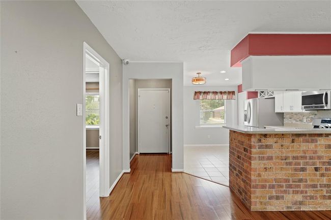 Photo showcases a welcoming entryway leading to a kitchen that opens to the living area. The space features vinyl wood flooring, a brick accented wall on the kitchen counter, modern stainless steel appliances, and ample natural light from large windows. | Image 4