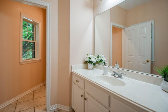 This bright bathroom features a large mirror, elegant countertop with a sink, and ample cabinet space. Natural light streams in from a window, highlighting the neutral walls and tiled floor. | Image 35