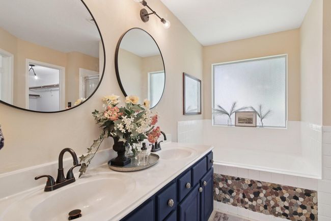 This bathroom features a dual sink vanity with elegant black fixtures and large round mirrors. The deep blue cabinetry complements the stone-accented tub area, creating a stylish and inviting space. Frosted windows provide privacy while allowing natural light. | Image 22