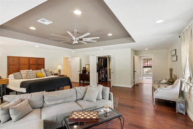 This spacious master bedroom features a tray ceiling with recessed lighting and a ceiling fan. It includes a large seating area, a cozy bed with stylish cushions, and direct access to a walk-in closet. The room is well-lit with natural light from large windows and has a warm, inviting hardwood floor. | Image 25