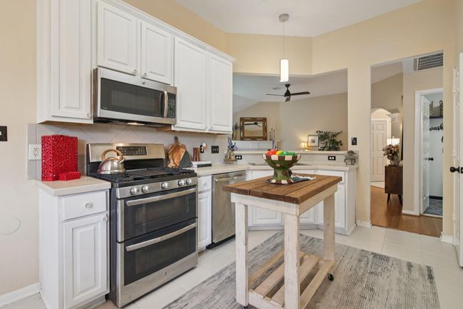 Bright kitchen with modern GE Profile stainless steel appliances and white cabinetry. It features a tile backsplash, pendant lighting, and an open layout connecting to a living area with hardwood flooring. | Image 12