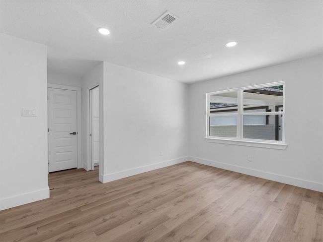 This photo showcases a bright, unfurnished room with light wood flooring, recessed lighting, and a large window offering natural light. The room features a white door and a clean, modern aesthetic. | Image 17