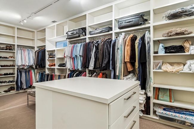 The PRIMARY CLOSET features a dual entry door, carpeted flooring, packing station with drawers, track lighting, raised privacy window, clothing compartments, built-in shelving, built-in hamper. | Image 25