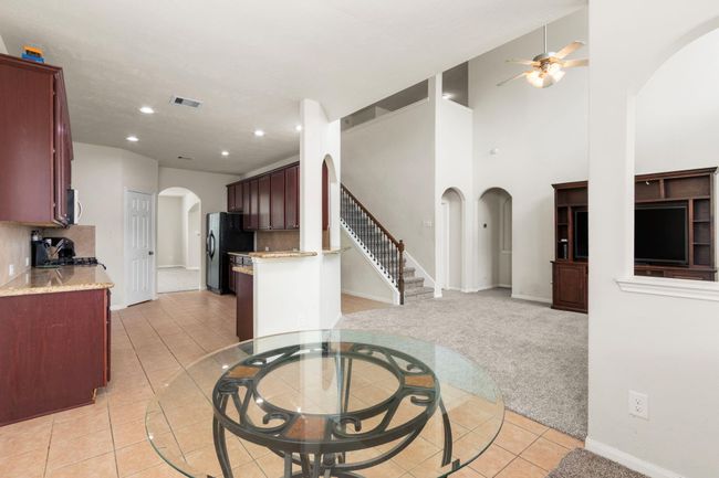 Spacious open-concept living area with high ceilings and a ceiling fan. The kitchen features dark wood cabinets, granite countertops, and tile flooring. A carpeted staircase leads to the upper level, and there is a built-in entertainment unit in the living room. | Image 11