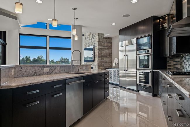 Kitchen with dark cabinets, exhaust hood, decorative light fixtures, light stone counters, and appliances with stainless steel finishes | Image 12