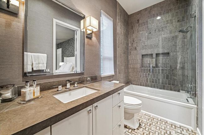 EN SUITE BATH features decorative tile flooring, papered walls, stone counter/backsplash, window with shade, tub/shower with subway tile surround/glass half partition, Kohler sink with decorative fixtures/cabinets and drawers below, metal framed mirror with 2 wall mount light sconces and walk-in closet. | Image 31