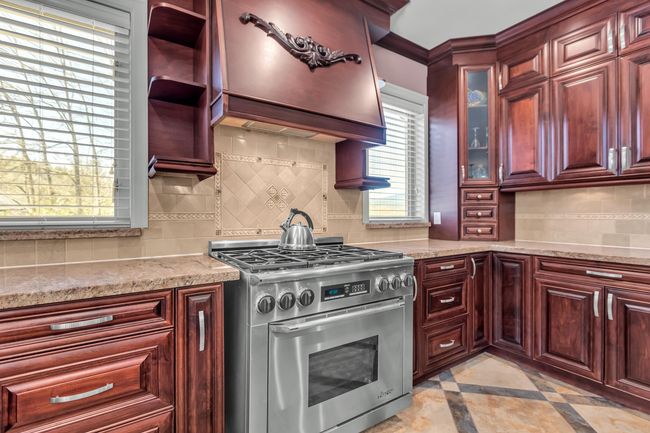 Kitchen with stainless steel gas range, reddish brown cabinets, backsplash, inlaid floor details, and glass insert cabinets | Image 14