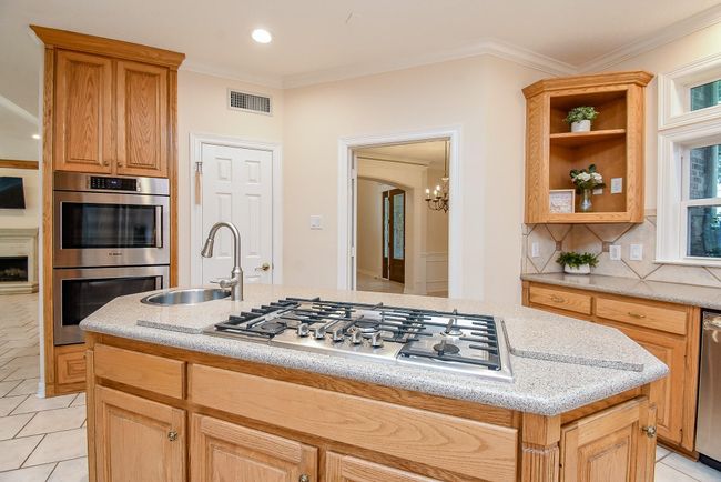 This kitchen features a central island with a gas cooktop and sink, surrounded by warm wooden cabinetry. Stainless steel double ovens are built into the cabinetry, and there's a view into an adjacent room with a chandelier. Natural light fills the space, highlighting the tiled backsplash and countertop. | Image 16