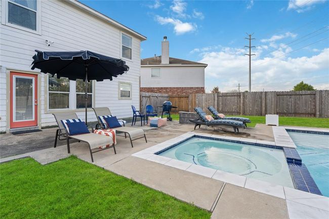 This backyard features a spacious patio with comfortable lounge chairs, a large umbrella for shade, and a sparkling pool with a hot tub, perfect for relaxation and entertaining. The area is enclosed by a privacy fence, offering a serene outdoor retreat. | Image 4