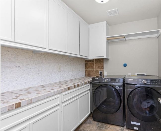 This is a utility space featuring modern, black front-loading washer and dryer units, with white cabinetry above for storage and a mosaic tile backsplash. The countertops are tiled, complementing the backsplash design. | Image 24