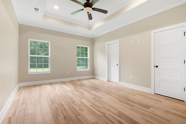 This room features light wood flooring, neutral walls, and a tray ceiling with recessed lighting and a ceiling fan. It has two large windows offering natural light and two white doors for access. | Image 13