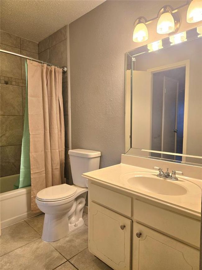 This bathroom features a clean, modern design with neutral tones. It includes a bathtub with a tiled shower, a white toilet, and a vanity with storage. The large mirror and overhead lighting add brightness to the space. | Image 13