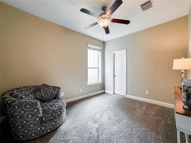 This first floor bedroom features neutral walls and carpet, a ceiling fan with light, and a cozy armchair. It has a window for natural light and a door leading to an adjoining space. Perfect for a small living area or office. | Image 16