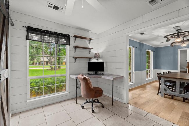 This photo showcases a bright home office space with large windows, providing ample natural light and views of the front yard. It features a corner desk, open shelves, and tile flooring. Adjacent is the dining, creating a seamless flow to the kitchen. | Image 8