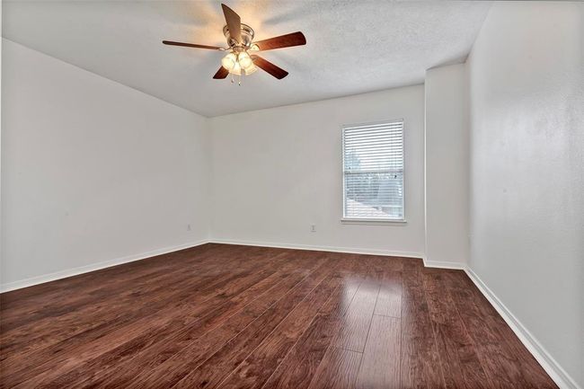 This room features clean white walls, rich wood flooring, a ceiling fan with lights, and a window allowing natural light. Perfect for a cozy, modern living space. | Image 27