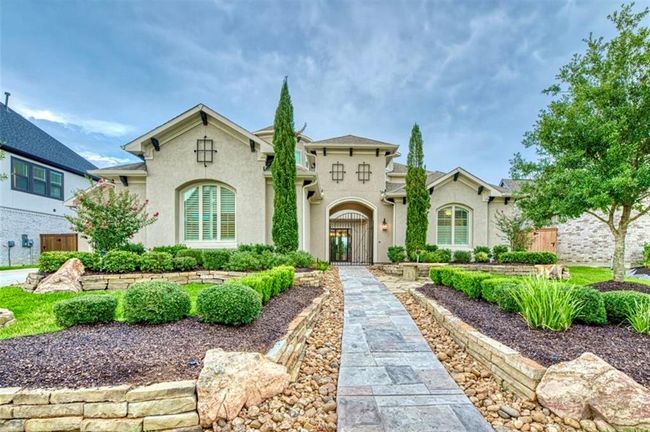 This stunning home features a welcoming stone pathway leading to an elegant entrance, flanked by lush landscaping with manicured shrubs and trees. The facade showcases a classic design with large windows and a neutral color palette, exuding charm and curb appeal. | Image 5