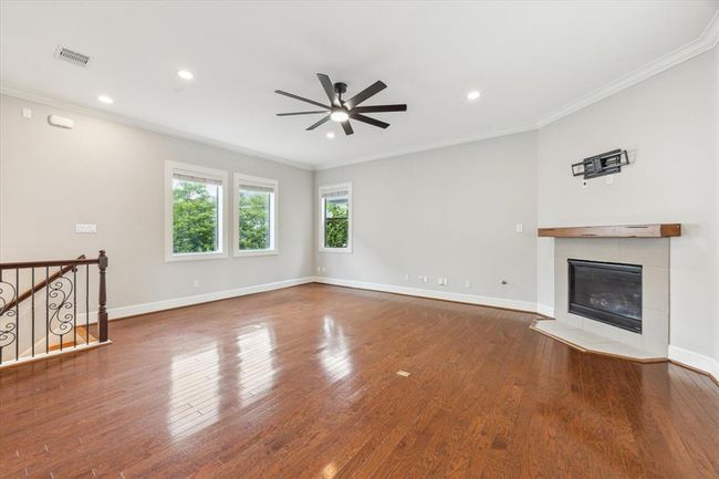 This spacious living room features hardwood floors, a modern ceiling fan, and a corner fireplace with a wooden mantle. Large windows provide natural light, and there's a stairway with decorative railing. Ideal for cozy gatherings and relaxation. | Image 9