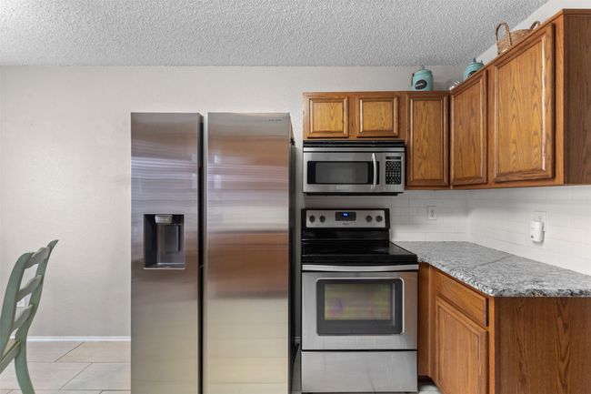 Kitchen with stainless steel appliances, a textured ceiling, brown cabinetry, light tile patterned floors, and decorative backsplash | Image 14