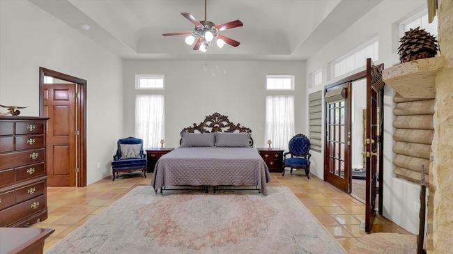 This is the second primary Bedroom featuring ceiling fan, french doors, a tray ceiling, and light saltillo floors, The room also has a corner stone and brick fireplace | Image 30