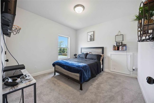 This bedroom features a cozy setup with a plush carpet, neutral walls, and a ceiling light. It includes a bed with a dark headboard, a wall-mounted TV, and a window offering natural light and outdoor views. Shelving adds extra storage and decor space. | Image 30
