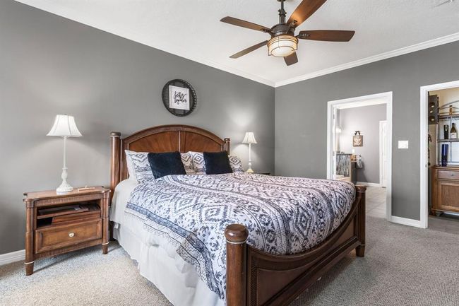 This cozy bedroom features a stylish gray wall color, a wooden bed frame, and matching nightstands. It has a ceiling fan, soft carpeting, and ample natural light from adjacent spaces. The room connects seamlessly to a bathroom, enhancing convenience and functionality. | Image 20