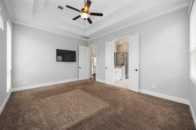 Bright bedroom with neutral walls, featuring plush carpet, a ceiling fan, and an en-suite bathroom with a double door entry. Ample natural light from multiple windows enhances the spacious feel. | Image 21