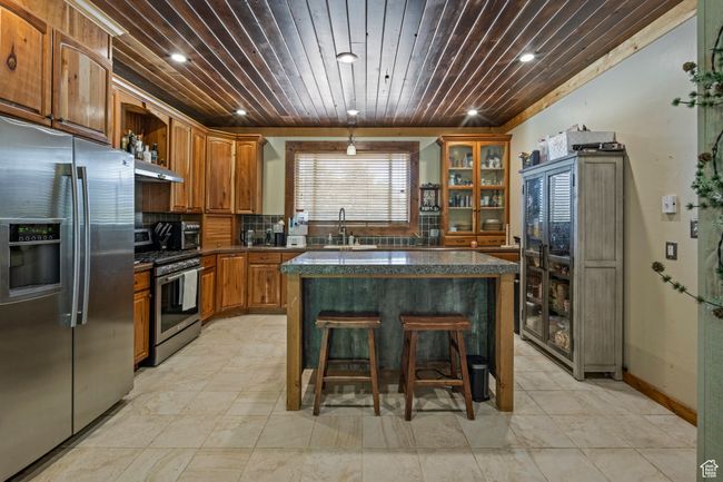 Kitchen featuring stainless steel appliances, brown cabinetry, under cabinet range hood, open shelves, and a kitchen bar | Image 8