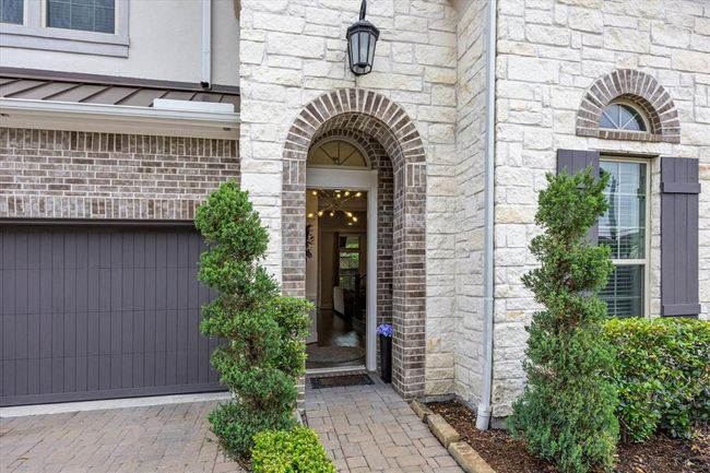 Charming entrance with a stone facade and arched doorway, flanked by well-maintained shrubs. The brick driveway leads to a modern garage, enhancing curb appeal. | Image 4