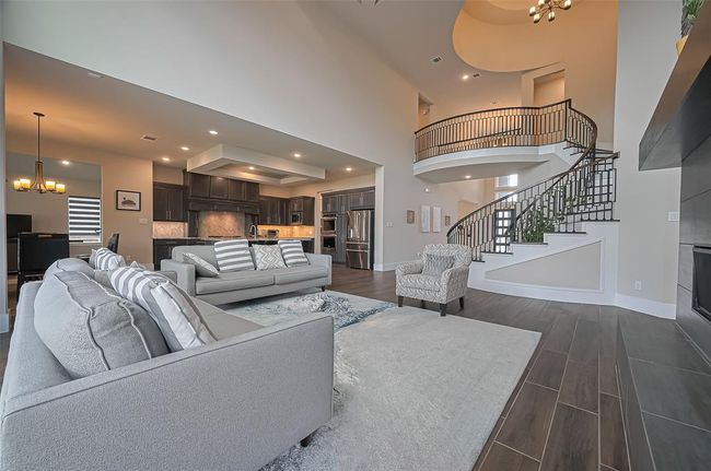 This spacious living area features an open-concept design with high ceilings, elegant wood tile flooring, and a sweeping staircase. The modern kitchen includes dark cabinetry and sleek finishes, while large windows provide ample natural light. A cozy fireplace and stylish furnishings complete the inviting atmosphere. | Image 10