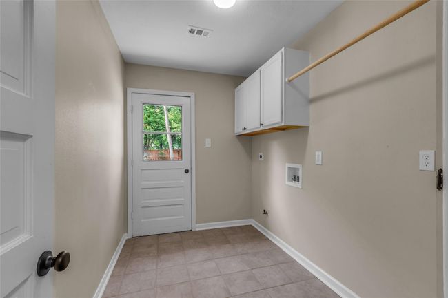 This is a utility or laundry room with neutral walls and tile flooring. It features a white overhead cabinet and broom closet for storage, hookups for a washer and dryer, and a back door with a window offering natural light. | Image 35