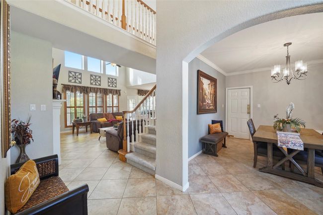 This photo showcases a bright, open-concept living area with high ceilings and ample natural light. To the left is a cozy living room with a fireplace, leading to a staircase. To the right, there's a formal dining area with elegant chandelier lighting and a welcoming entryway. The space features neutral tones and tile flooring, providing a warm and inviting atmosphere. | Image 9