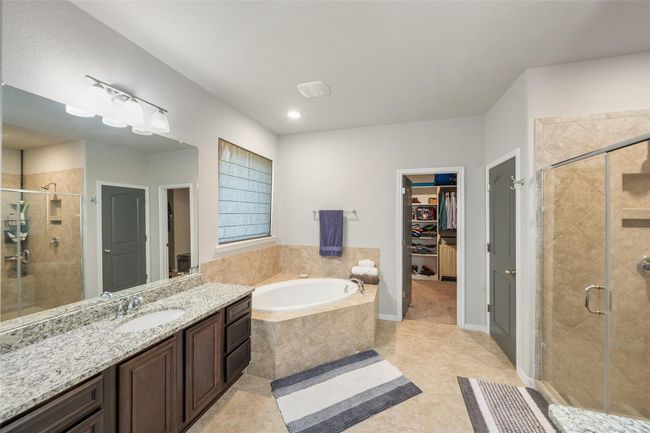 The primary bathroom features a luxurious soaking tub, a large glass-enclosed shower, and a dual-sink vanity with granite countertops. It offers ample storage and connects to a walk-in closet, creating a practical and elegant space. | Image 18