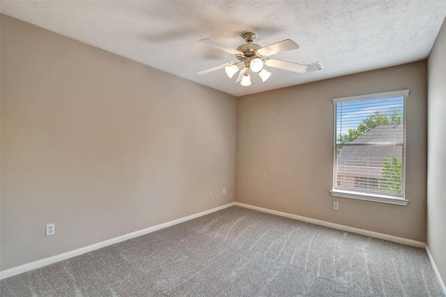 This is a cozy, neutral-toned bedroom featuring carpeted floors, a ceiling fan with lights, and a window that offers natural light and a view of the outside. | Image 38