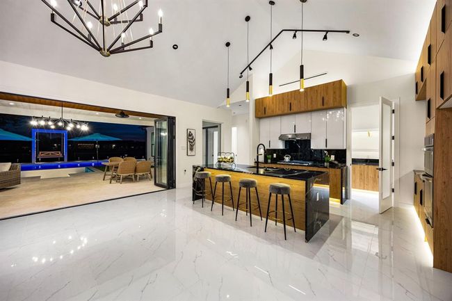This breathtaking kitchen offers a seamless blend of modern luxury and functionality, highlighted by dramatic black granite countertops and a waterfall island. High-gloss cabinetry paired with warm wood creates a striking contrast, while pendant and track lighting add a sophisticated ambiance. Expansive sliding doors open to a resort-style outdoor space with pool views, enhancing the indoor-outdoor living experience. Gleaming marble floors and clean architectural lines complete the contemporary design. | Image 10