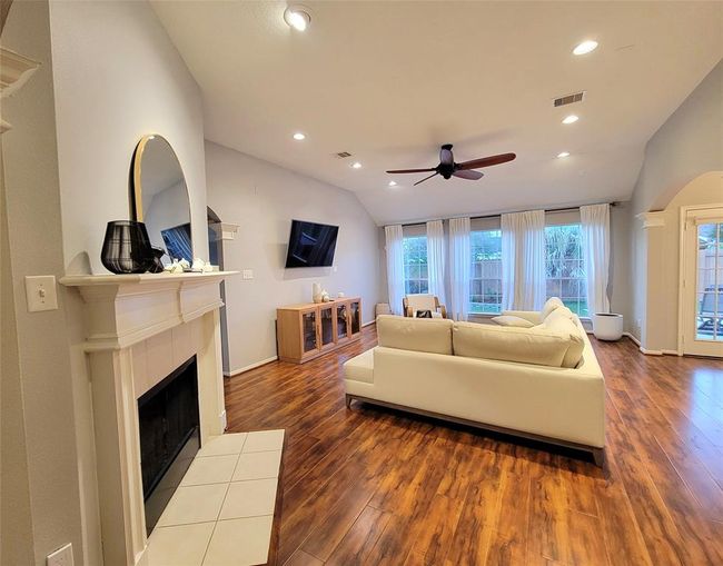 Spacious living room with warm wood flooring, a cozy fireplace, and large windows offering plenty of natural light. It features a modern ceiling fan, elegant decor, and a comfortable seating area. | Image 12