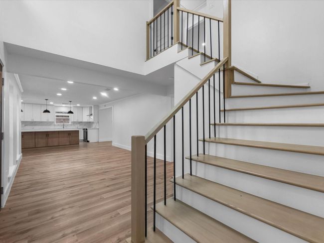 This photo showcases a modern, open-concept living space with sleek wooden flooring and a stylish staircase. The kitchen features elegant white cabinetry, marble countertops, and pendant lighting, creating a bright and inviting atmosphere. | Image 4