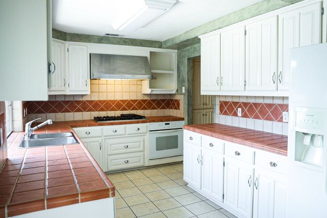 Kitchen with white appliances, tile counters, wall chimney exhaust hood, light tile patterned floors, and white cabinets | Image 13