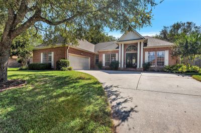 This is a beautiful single-story brick home featuring a two-car garage, a prominent entryway with a peaked roof, large windows, and a well-maintained lawn with mature trees. | Image 1