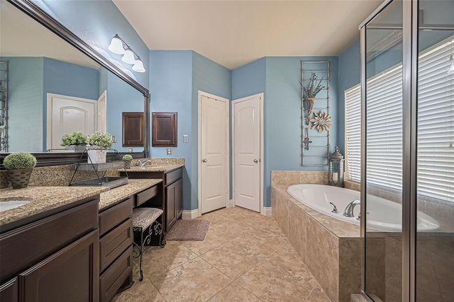 The ensuite bathroom impresses with elegant designer paint, calming neutral tile floors, and a large vanity complete with dual sinks and a comfortable seating space. | Image 23