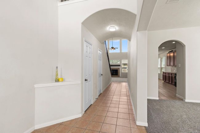 This photo showcases a spacious hallway with high ceilings, leading to a living area featuring large windows and a fireplace. To the right, there's an arched entry to a kitchen with ample cabinet space. The flooring is a mix of tile and carpet, creating a warm, inviting atmosphere. | Image 5