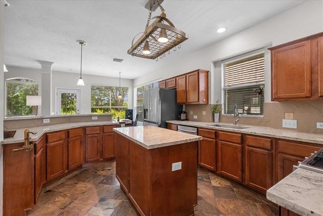 Spacious kitchen with rich wooden cabinetry, granite countertops, and a central island. Features stainless steel appliances, large windows for natural light, and elegant overhead lighting with custom hanging pot rack. | Image 8